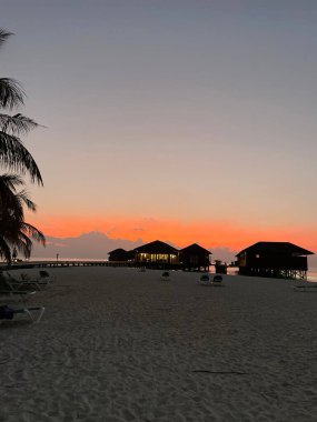 Sahildeki küçük kulübelerin dikey bir görüntüsü. Canlı bir günbatımı arka planında.