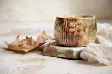 Ahşap bir yüzey üzerinde eski bir seramik kupanın yakın plan bir fotoğrafı. Yanında bez ve kurumuş bir yaprak var.
