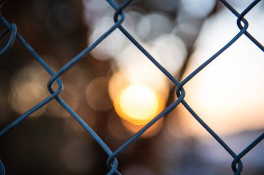 A closeup of a metal chain fence with beautiful orange and white bokeh lights visible through the gaps in the background