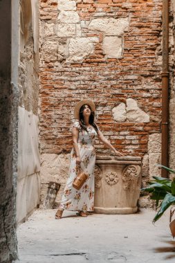 A vertical shot of a Caucasian woman with black hair in a floral dress and a hat posing near ruins