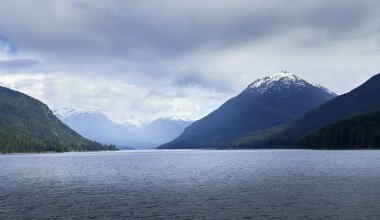 Bulutlu bir günde, önplanda sakin bir göl olan uzak, engebeli, karla kaplı dağ tepeleri.