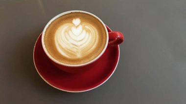 A top view of a delicious hot latte in a red cup with a heart design made with foam on a gray table