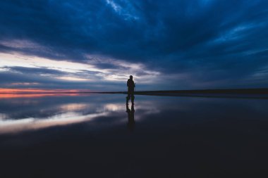 A scenic view of a sunset with a man's silhouette on a frozen lake with reflection