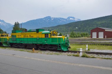 An industrial green and yellow Yukon train rides next to an asphalt road in Alaska, USA