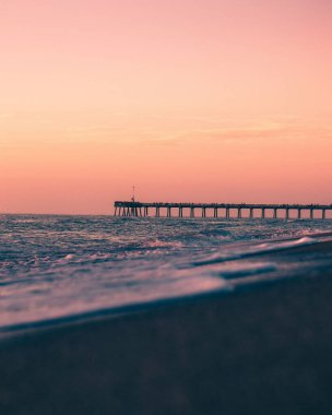 A vertical distant view of a pier in the ocean with sunset in the background