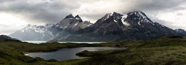 A panoramic shot of lake against a landscape covered with greenery and snowy mountains on a gloomy day