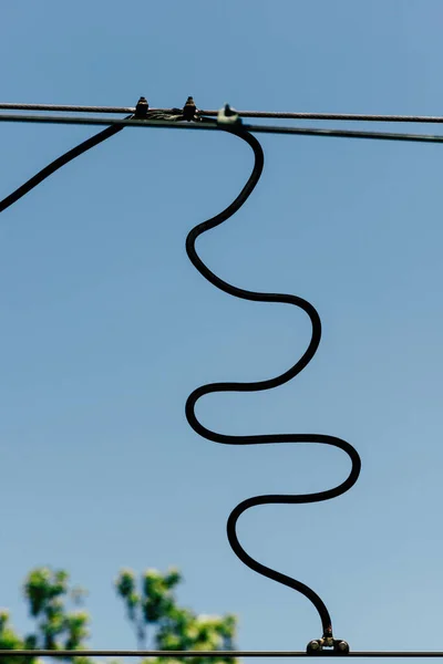 A closeup shot of a jumper wire connecting the single catenary wire to the dual contact wires against blue sky on a sunny day with blurred background