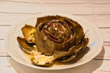 Italian style boiled artichoke on white plate on wooden table stuffed with egg, parsley, garlic , typical mediterranean food, closeup