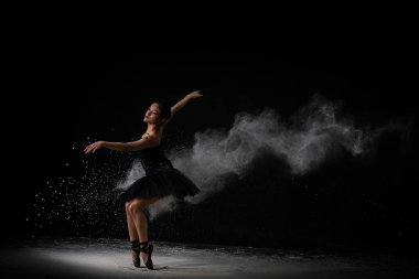 A breathtaking shot of a ballerina in black clothes dancing in a dark Studio in a cloud of dust