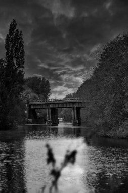 A black and white vertical shot of bridge over a river with clouds in the sky and reflection in water