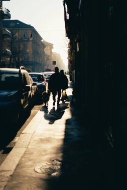 A vertical shot of people's silhouettes walking by the street between buildings and cars