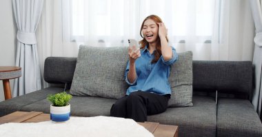 Evde akıllı telefonlu Asyalı genç bir kadının portresi. Çekici kız kanepede oturmuş, rahat bir yerde arkadaşlarıyla sohbet ediyor.