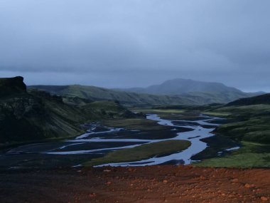 A scenic view of rivers flowing through a green mountain range against a cloudy sky in Iceland