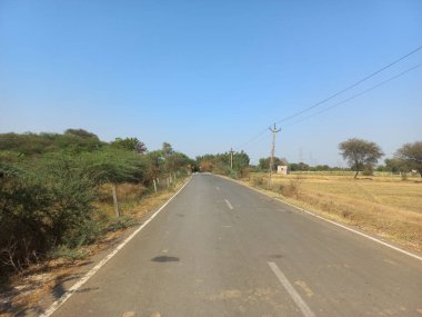 A beautiful shot of an asphalt road with trees on a side and farmland on other side under clear sky