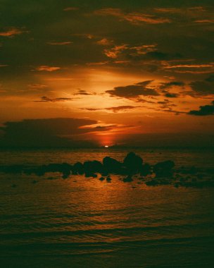 Gün batımında bir deniz manzarasının dikey görüntüsü. Suda kaya silueti var.