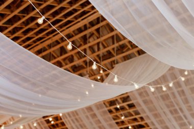 A beautiful shot of white curtains hanging on a wooden suspended ceiling with yellow lights, a wedding decor concept
