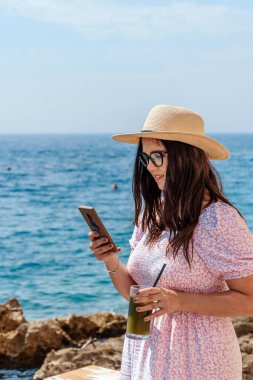 Bir kadının telefonunu tutarken ve sahilde lezzetli bir içkinin keyfini çıkarırken çekilmiş dikey bir fotoğrafı.