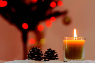 A closeup shot of a glass candle and two pine cones on a snowflakes with blurred red background