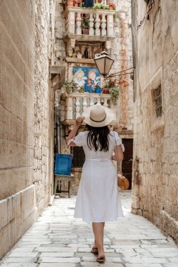 A vertical closeup of a young woman in a white dress walking along a narrow street in Trogir, Croatia.