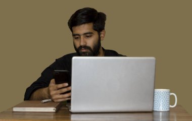 A South Asian man looking at his phone while working on a laptop