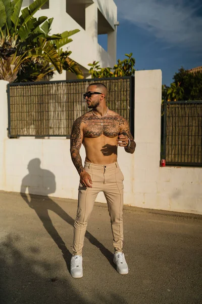 A vertical shot of a young tattooed shirtless man in beige pants and white sneakers
