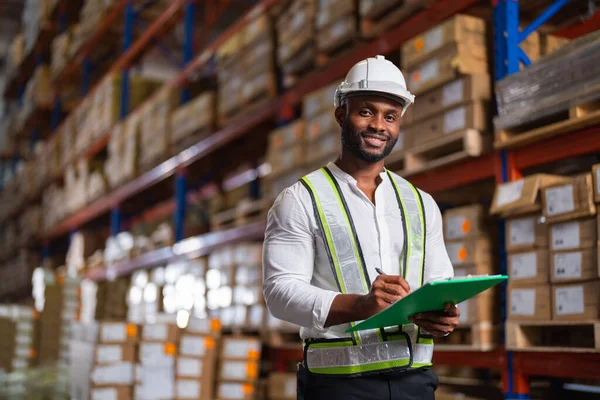 A portrait of a black warehouse manager holding a clipboard checking inventory in a large distribution center