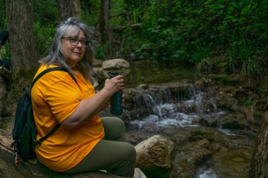 older white-haired woman drinking water from a bottle in a field with a waterfall in the background