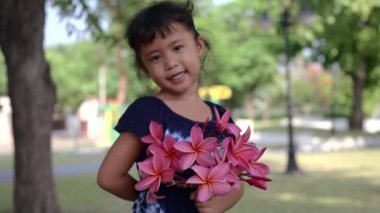 A closeup shot of an Asian little girl holding pink flowers and showing them to the camera with a smile on her face on a sunny day