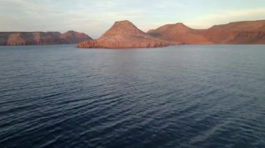 An aerial view of the wavy sea and the rocky mountains gleaming under the cloudy blue sky in 4K
