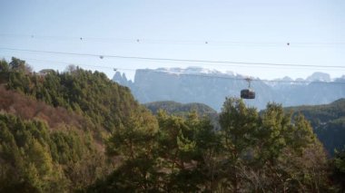 A cable car over the dense forests surrounded by snow-capped rocky mountains in Bolzano, Dolomites