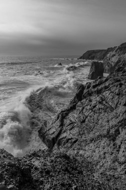 A vertical black and white shot of coastal waves in cloudy sky background