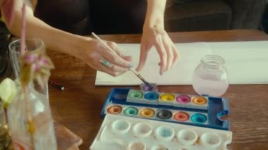 A closeup shot of a female hand drawing on a white paper with a painting brush near palette of colors on a wooden table