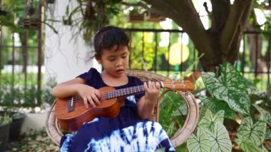 A Little cute Southeast Asian girl playing ukulele and singing in the garden