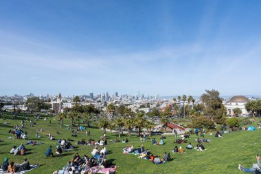A wide-angle photo of crowds of people gathered at Mission Dolores Park with the San Francisco skyline in the background.