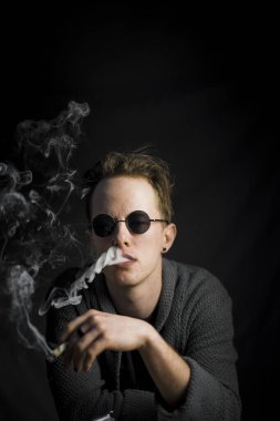 The vertical shot of a smoking young guy wearing round black sunglasses isolated on the black background