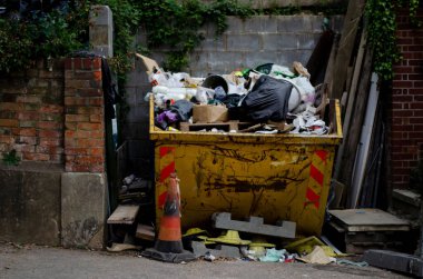 A full yellow rubbish skip full of trash from a building site. Wood and other building materials around it.  Ready to go to the dump.