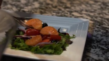 A professional chef adding mandarins, tomatoes and onions to salad greens on a white plate