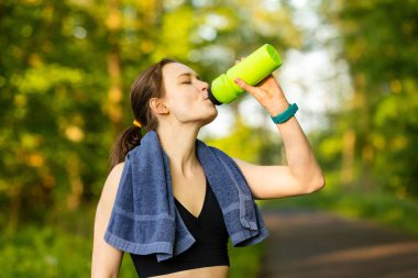 A young Caucasian female with a towel drinking from a bottle outside in the forest