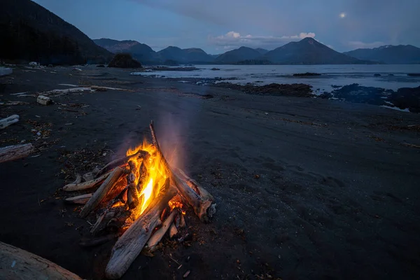 A campfire on calm lake shore with mountains in the background