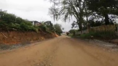 An unpaved road with green trees, and traditional rural houses on the sides of a road in daylight