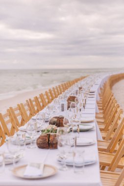 A long table with wooden chairs and glass cups, plates with cake by the sea under cloudy sky