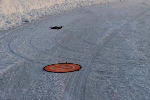The view of the flying drone and an orange landing pad on the ground covered with snow.