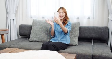 Evde akıllı telefonlu Asyalı genç bir kadının portresi. Çekici kız kanepede oturmuş, rahat bir yerde arkadaşlarıyla sohbet ediyor.