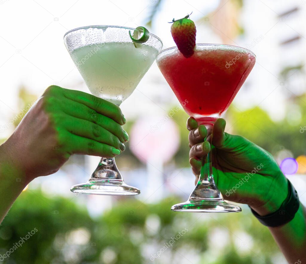 A closeup of two hands cheering with a strawberry mocktail and a blended margarita cocktail