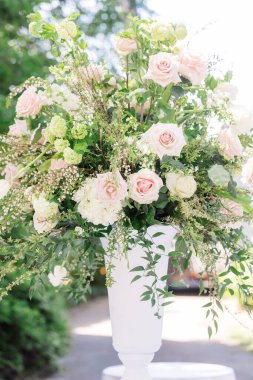 A vertical shot of a composition of white roses, hydrangeas and green branches in a white vase under the rays of the sun
