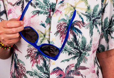 A man holding a blue sunglasses wearing tropical shirt
