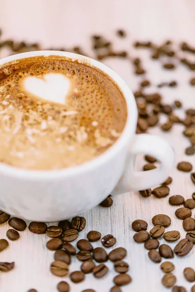 A vertical, close-up shot of a cup of coffee with a heart foam surrounded by coffee beans