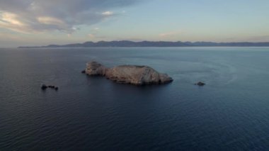 An aerial view of a rocky island in the wavy sea under the cloudy blue sky in 4K
