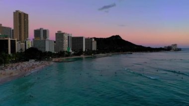 A drone shot of sunset over buildings and skyscrapers on a beautiful beach with crystal water under cloudy sky in Waikiki, Hawaii