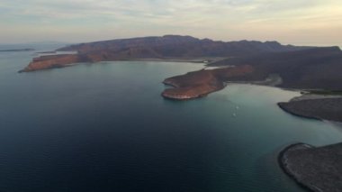 An aerial view of the wavy sea and the rocky mountains gleaming under the cloudy blue sky in 4K